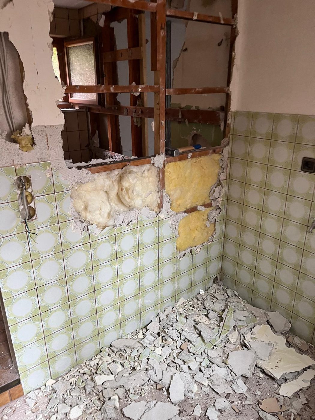 Altes Bad im Rückbau: grün gemusterte Wandfliesen, geöffnete Ständerwand mit Dämmwolle, Fliesenschutt auf dem Boden