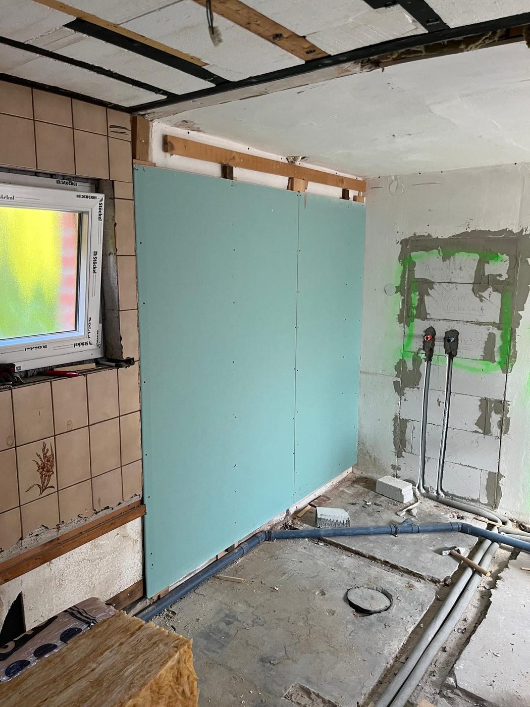 Neue Vorwand aus grüner Feuchtraum-Gipsplatte, daneben Betonwand mit angezeichneten Anschlüssen und neu verlegten Wasserleitungen