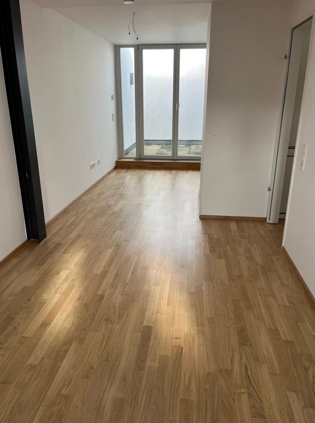 Leerer Wohnraum mit frisch versiegeltem Eichenparkett, weiße Wände und bodentiefe Fenstertür zur Terrasse